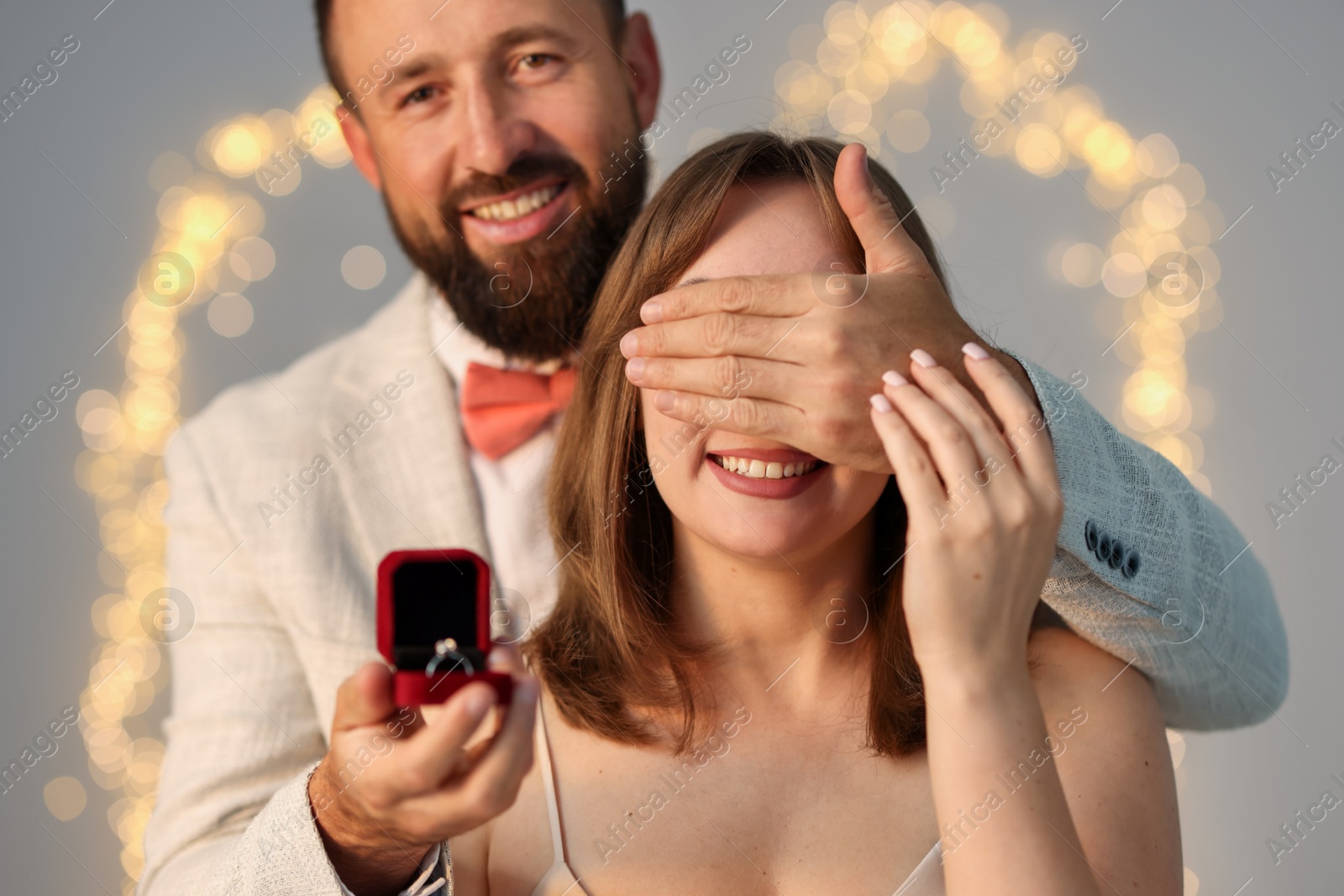 Photo of Man with engagement ring making marriage proposal to his girlfriend on light grey background with blurred lights, bokeh effect