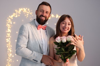Photo of Making marriage proposal. Woman with engagement ring and her happy fiance on light grey background with blurred lights, bokeh effect