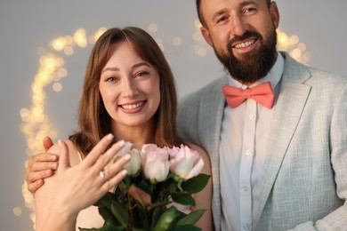Photo of Making marriage proposal. Woman with engagement ring and her happy fiance on light grey background with blurred lights, bokeh effect
