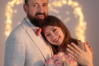 Photo of Making marriage proposal. Woman with engagement ring and her happy fiance on light grey background with blurred lights, bokeh effect