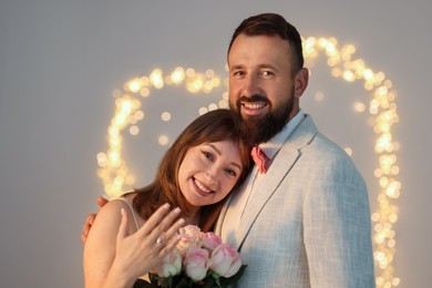 Photo of Making marriage proposal. Woman with engagement ring and her happy fiance on light grey background with blurred lights, bokeh effect