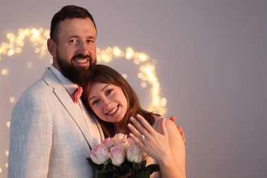Photo of Making marriage proposal. Woman with engagement ring and her happy fiance on light grey background with blurred lights, space for text. Bokeh effect