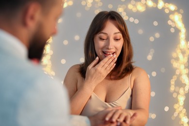 Photo of Making marriage proposal. Man putting engagement ring on his girlfriend's finger against light grey background, closeup. Bokeh effect