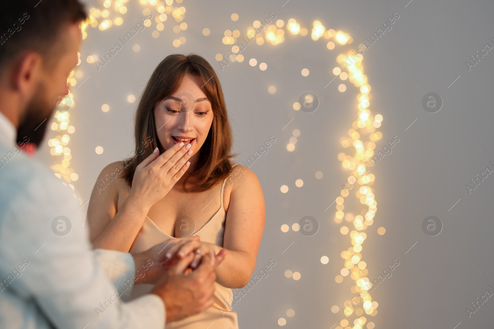 Photo of Making marriage proposal. Man putting engagement ring on his girlfriend's finger against light grey background, space for text. Bokeh effect