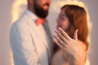 Photo of Making marriage proposal. Woman showing engagement ring and hugging her fiance on light grey background with blurred lights, selective focus. Bokeh effect