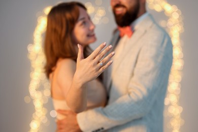 Photo of Making marriage proposal. Woman showing engagement ring and hugging her fiance on light grey background with blurred lights, selective focus. Bokeh effect