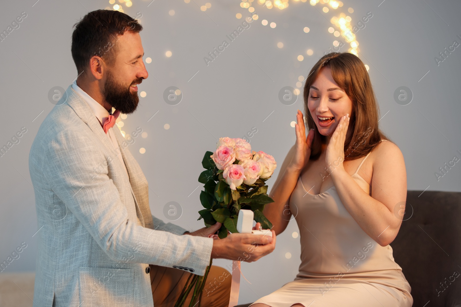 Photo of Man with engagement ring and bouquet of flowers making marriage proposal to his girlfriend on light grey background, bokeh effect