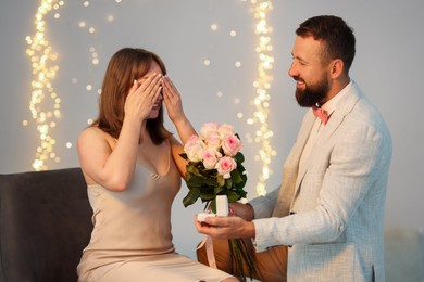 Photo of Man with engagement ring and bouquet of flowers making marriage proposal to his girlfriend on light grey background, bokeh effect