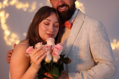 Photo of Making marriage proposal. Woman with engagement ring, flowers and her fiance on light grey background, bokeh effect
