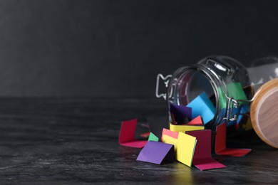 Photo of Colorful paper notes in glass jar on black table, closeup. Space for text