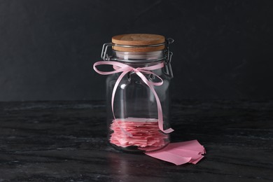 Photo of Pink paper notes in glass jar with ribbon on black table