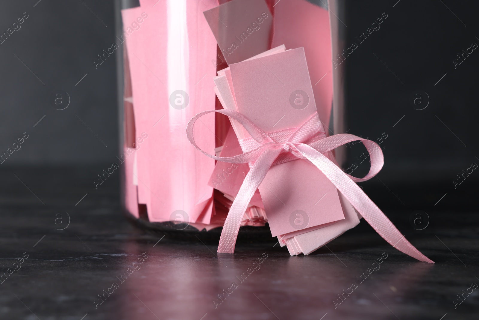 Photo of Pink paper notes in glass jar with ribbon on black table, closeup