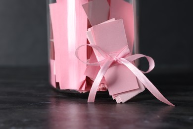 Photo of Pink paper notes in glass jar with ribbon on black table, closeup