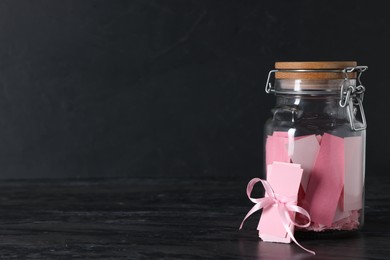 Photo of Pink paper notes in glass jar with ribbon on black table, space for text
