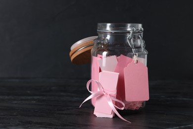 Photo of Pink paper notes in glass jar with ribbon on black table, space for text