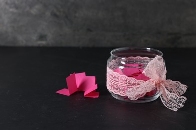 Photo of Pink paper notes in glass jar with ribbon on black table, space for text