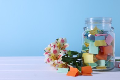 Photo of Colorful paper pieces in glass jar and flowers on white wooden table against light blue background, closeup. Space for text