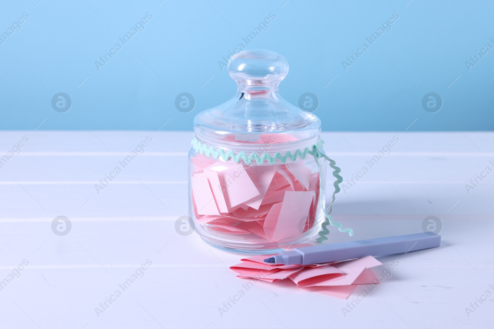 Pink paper pieces in glass jar and marker on white wooden table against light blue background, closeup Photo of Pink paper pieces in glass jar and marker on white wooden table against light blue background, closeup