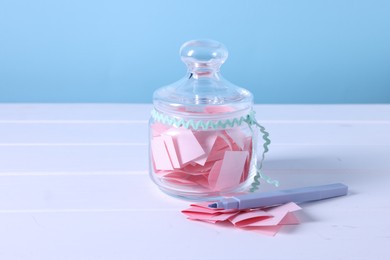 Photo of Pink paper pieces in glass jar and marker on white wooden table against light blue background, closeup