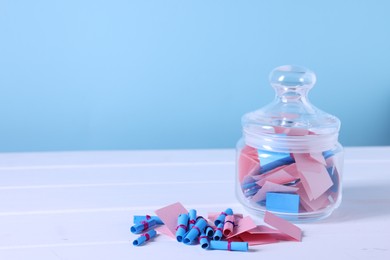 Photo of Colorful paper pieces in glass jar on white wooden table against light blue background, closeup. Space for text