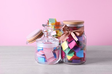 Photo of Colorful paper pieces in glass jars on wooden table against pink background. Message notes