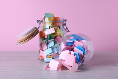 Photo of Colorful paper pieces in glass jars on wooden table against pink background, closeup. Message notes