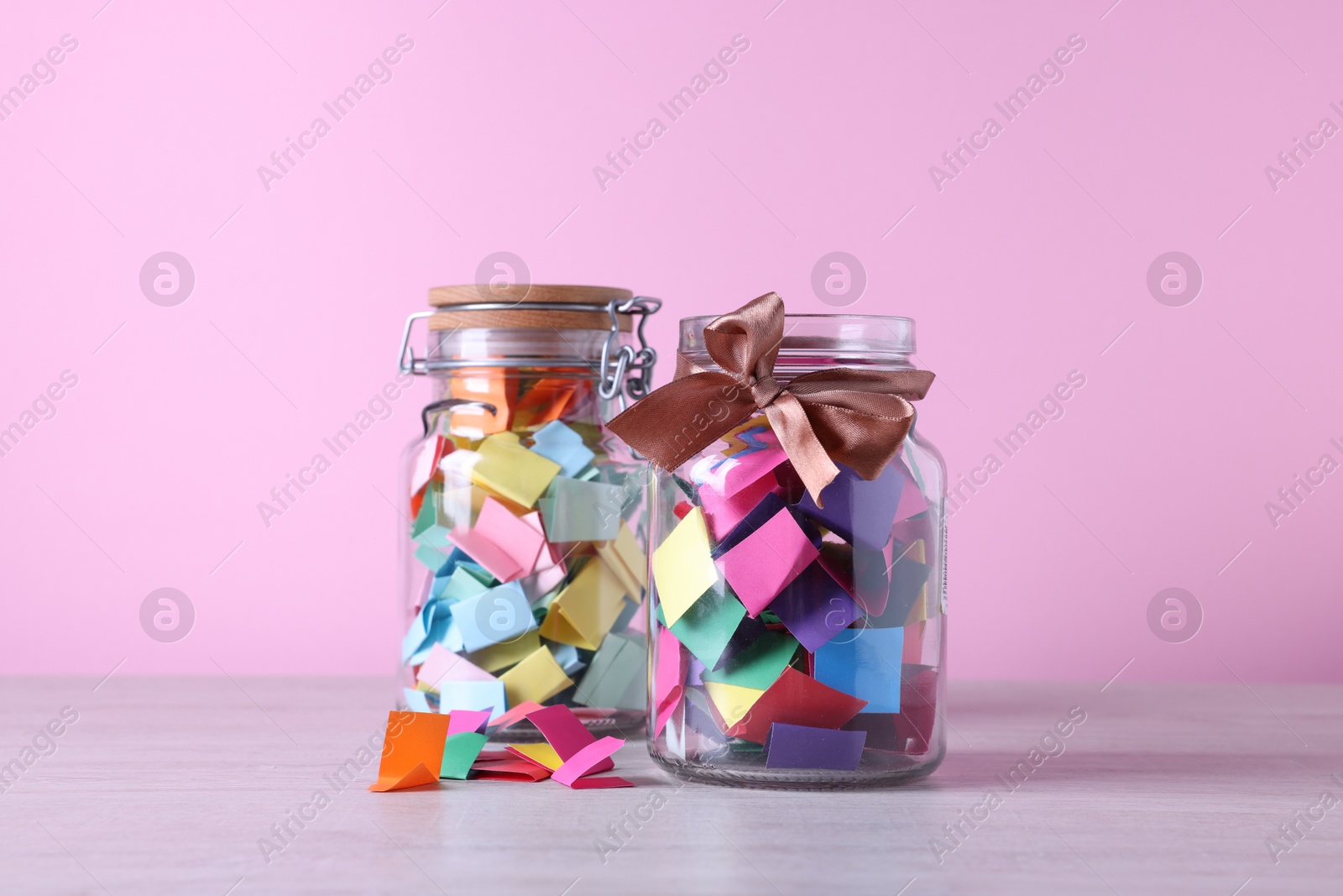 Colorful paper pieces in glass jars on wooden table against pink background. Message notes Photo of Colorful paper pieces in glass jars on wooden table against pink background. Message notes