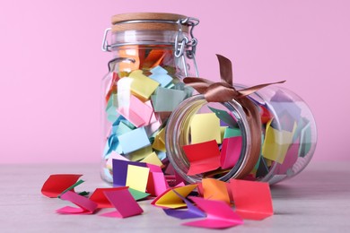 Photo of Colorful paper pieces in glass jars on wooden table against pink background, closeup. Message notes