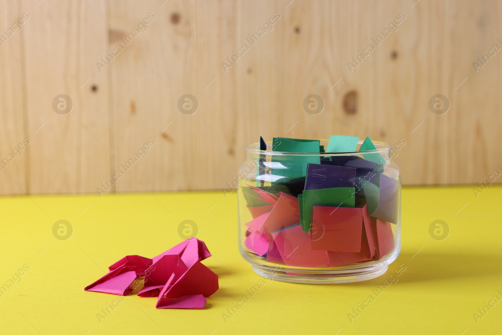 Colorful paper notes in glass jar on yellow table against wooden background, closeup Photo of Colorful paper notes in glass jar on yellow table against wooden background, closeup