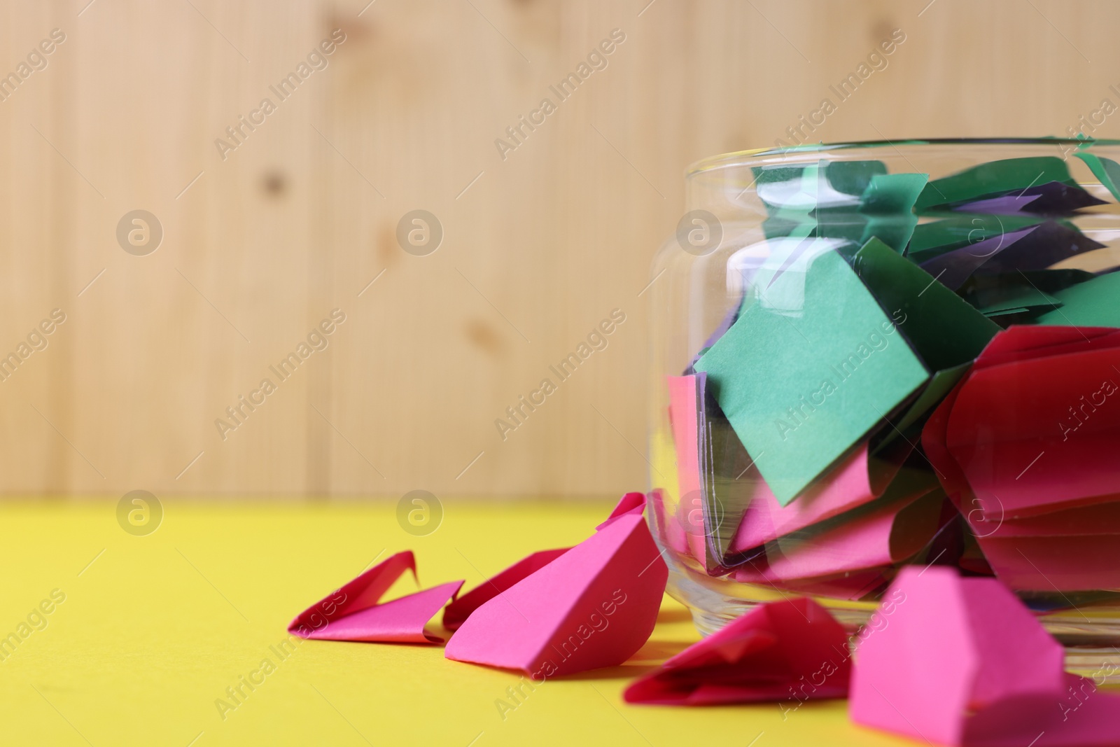 Colorful paper notes in glass jar on yellow table against wooden background, closeup. Space for text Photo of Colorful paper notes in glass jar on yellow table against wooden background, closeup. Space for text