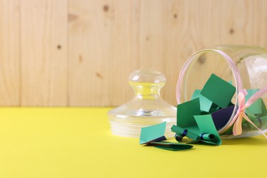 Photo of Colorful paper notes in glass jar on yellow table against wooden background, closeup. Space for text
