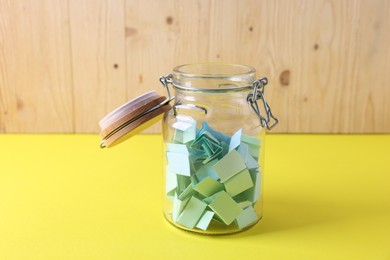 Photo of Colorful paper notes in glass jar on yellow table against wooden background