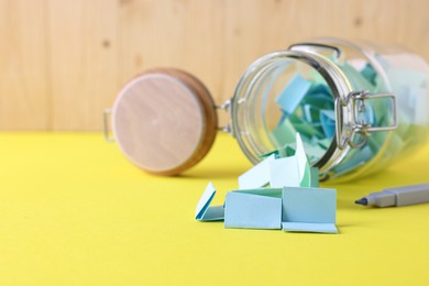 Photo of Colorful paper notes in glass jar and marker on yellow table, closeup. Space for text