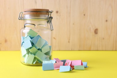 Photo of Colorful paper notes in glass jar on yellow table against wooden background