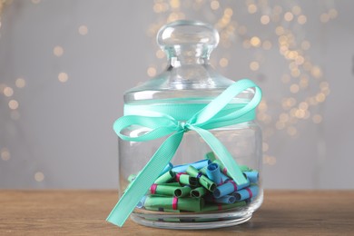 Photo of Colorful paper notes in glass jar on wooden table against grey background with blurred lights, closeup. Bokeh effect