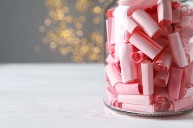 Photo of Pink paper notes in glass jar on white wooden table against grey background with blurred lights, space for text. Bokeh effect
