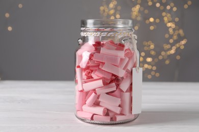 Photo of Pink paper notes in glass jar on white wooden table against grey background with blurred lights, closeup. Bokeh effect