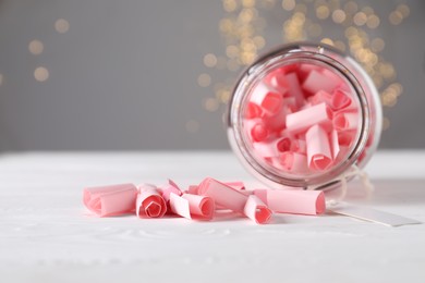 Photo of Pink paper notes in glass jar on white wooden table against grey background with blurred lights, closeup. Bokeh effect