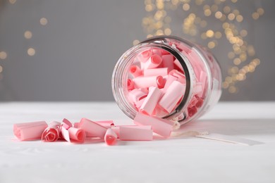 Photo of Pink paper notes in glass jar on white wooden table against grey background with blurred lights, closeup. Bokeh effect