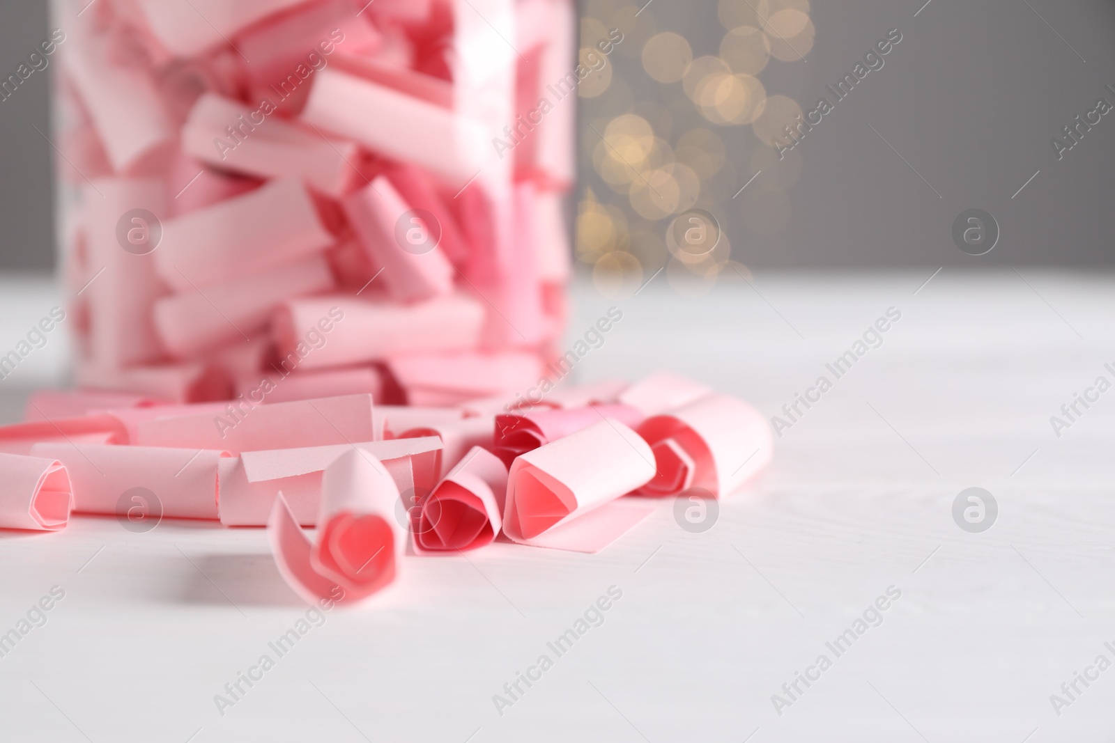 Photo of Pink paper notes in glass jar on white wooden table against grey background with blurred lights, space for text. Bokeh effect