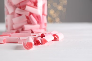 Photo of Pink paper notes in glass jar on white wooden table against grey background with blurred lights, space for text. Bokeh effect