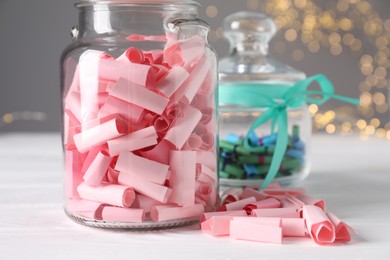 Photo of Colorful paper notes in glass jars on white wooden table against grey background with blurred lights, closeup. Bokeh effect