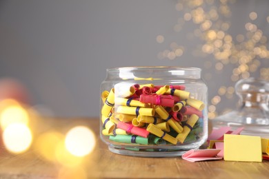 Photo of Colorful paper notes in glass jar on wooden table against grey background with blurred lights, space for text. Bokeh effect