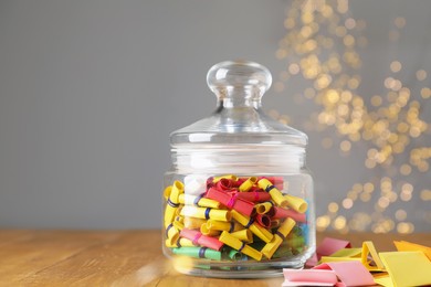 Photo of Colorful paper notes in glass jar on wooden table against grey background with blurred lights, space for text. Bokeh effect