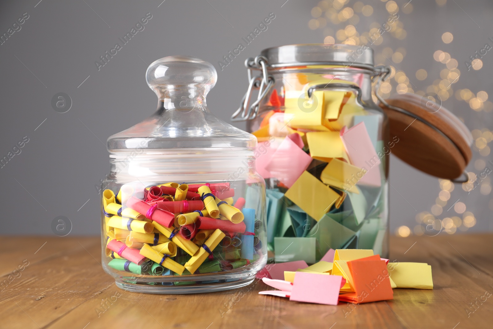 Colorful paper notes in glass jars on wooden table against grey background with blurred lights. Bokeh effect Photo of Colorful paper notes in glass jars on wooden table against grey background with blurred lights. Bokeh effect