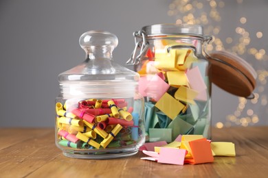 Photo of Colorful paper notes in glass jars on wooden table against grey background with blurred lights. Bokeh effect