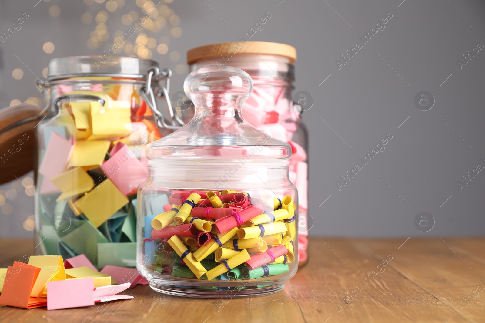 Colorful paper notes in glass jars on wooden table against grey background with blurred lights, space for text. Bokeh effect Photo of Colorful paper notes in glass jars on wooden table against grey background with blurred lights, space for text. Bokeh effect