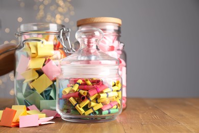 Photo of Colorful paper notes in glass jars on wooden table against grey background with blurred lights, space for text. Bokeh effect