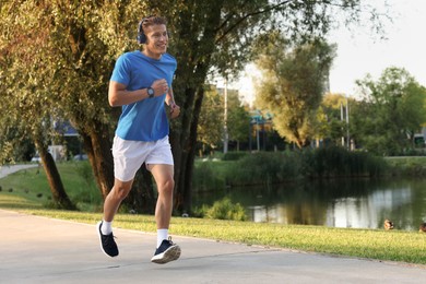 Photo of Happy man with headphones running near lake in park, space for text