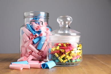 Photo of Colorful paper notes in glass jars on wooden table against grey background, closeup
